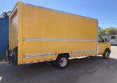 A large yellow delivery truck is parked on a paved area outdoors. The truck has a box-style cargo area with silver horizontal stripes. The sky is clear and blue, and a building with a blue wall is partly visible on the left.