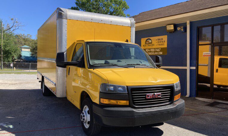 A yellow GMC box truck is parked outside a building with a blue and yellow exterior. The signage indicates sales, service, and finance options. Another similar truck is reflected in the building’s windows.