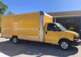 A bright yellow box truck is parked outside a building on a sunny day. The truck has a silver metallic top and is positioned on a paved surface with trees and a sign partially visible in the background.
