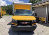 A yellow GMC box truck is parked in front of a building with a "Sales & Rentals" sign. Other yellow trucks are visible in the background under a partly cloudy sky.