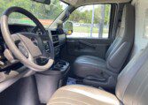 The interior of a GMC vehicle, showing the driver and passenger seats, steering wheel, dashboard, and center console with cup holders. Sunlight streams through the windows, revealing a view of a fence and trees outside.