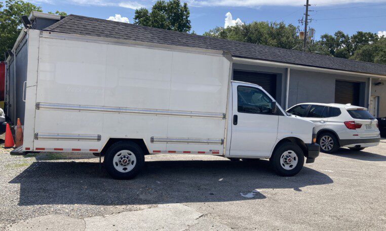 A white box truck is parked outside a gray building with garages, next to a white SUV, on a sunny day with trees and a blue sky in the background.