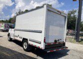 A white box truck is parked outdoors on a sunny day near a fence and other vehicles, with trees and a partly cloudy sky in the background.